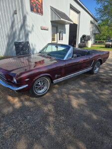 Classic burgundy Ford Mustang convertible parked outside an auto shop on a sunny day.