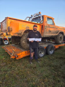Man standing in front of a muddy orange pickup truck on a trailer holding papers in a grassy outdoor area.